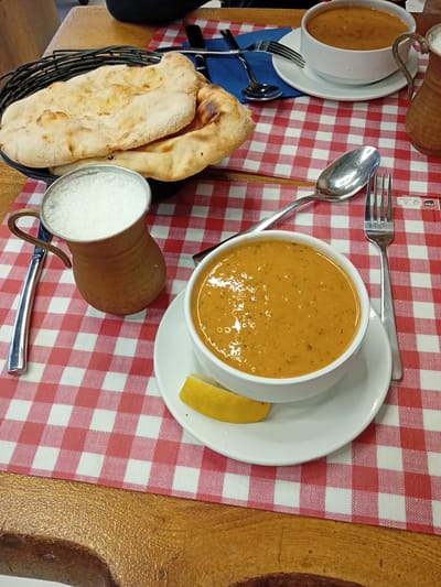 The image depicts a table setting, likely in a restaurant, featuring a checkered red and white tablecloth. A bowl of soup, accompanied by a lemon wedge and placed on a white saucer, sits next to a copper mug filled with a foamy beverage. A knife, fork, and spoon are arranged nearby. A basket containing flatbread is positioned on the left side of the table, near another bowl of soup and cutlery. The table surface appears to be wooden. The image does not provide any identifying features that indicate the location is Cenon, France.