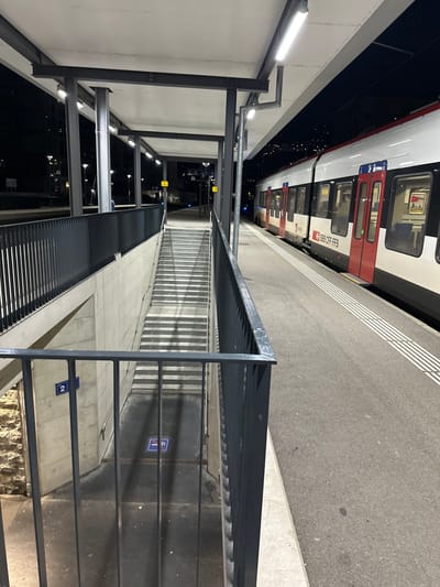 The image depicts a night scene at a train station platform in Vevey, Switzerland. A white and red SBB CFF FFS train is stationary on the right side of a grey asphalt platform, featuring white tactile paving lines along its edge. The platform is covered by a grey canopy supported by dark grey steel pillars, illuminated by multiple overhead fluorescent tube lights. To the left, a concrete staircase with dark grey metal railings descends into an underpass. A blue sign bearing the number "2" is affixed to the concrete wall adjacent to the stairs, and a blue and white pedestrian symbol is visible on the ground at the staircase's base. The background beyond the platform is dark, displaying scattered distant city lights. No individuals are present in the scene.