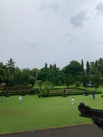 The image shows a view from a high vantage point looking down at a grassy courtyard with a stone wall bordering it. The courtyard is part of the Candi Sewu temple complex in the city of Mungkid, Indonesia. The coordinates -7.605, 110.23 confirm the location. It's a cloudy day. Two people are visible in the courtyard, one walking and one standing still. Several people in the distance are also visible. There are a variety of green trees and bushes around the courtyard. In the foreground, there is a stone structure that appears to be part of a temple. A few garbage cans are present in the courtyard. There are some other structures visible beyond the courtyard. The scene is relatively quiet and peaceful.