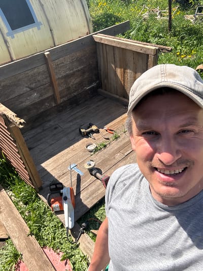 A selfie of a man wearing a gray t-shirt and a gray baseball cap. He is positioned above and to the right of a wooden structure being built outside in the grass in the city of Уром, Russia. On the wooden structure are various tools, including a cordless drill, a metal grinder, and a small chainsaw. There are also small green plants on the wood.