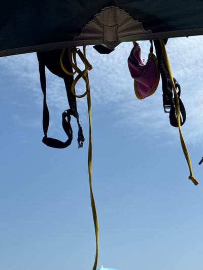 An upward-facing shot captures a bright blue sky with scattered clouds, as viewed from beneath a dark green tent. Several items are suspended from the tent's edge by yellow cords. On the left, a black strap with white lettering appears to be a part of a bag or backpack, with a small silver metal tag hanging from it. To the right of this, a pink bikini top is suspended, its fabric slightly ruffled. Further to the right, another black strap, possibly from the same bag as the first, is visible, connected to a complex system of black straps and buckles. A long, bright yellow cord dangles down from the tent, tied in a knot at its end, suggesting it might be part of a clothesline or a securing strap. The scene is set outdoors, possibly during a camping trip in or near the city of Gavà, Spain, given the bright, sunny weather.