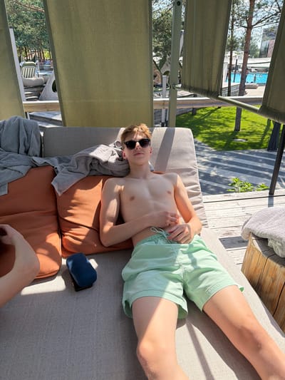 The image shows a young person lying on a lounge chair outdoors. The subject is shirtless, wearing light green shorts and sunglasses. They are lying on their back, with their hands resting on their stomach. The lounge chair is upholstered in gray fabric and has orange cushions. Two gray towels are draped over the chair. To the right of the chair, there is a small blue object, possibly a travel pillow.

In the background, we see a swimming pool with people in it, a grassy area with trees, and some buildings. The sun is shining brightly, casting shadows, suggesting it is a sunny day. The scene appears to be a resort or a residential area with a pool. The overall mood seems relaxed and summery.

The image is taken from a slightly elevated angle, looking down at the subject. The orientation of the image is rotated, with the subject's head towards the left and their feet towards the right. There is no visible text in the image. The location context of Khotianivka, Ukraine, combined with the presence of a pool and outdoor furniture, suggests a recreational setting during warm weather.