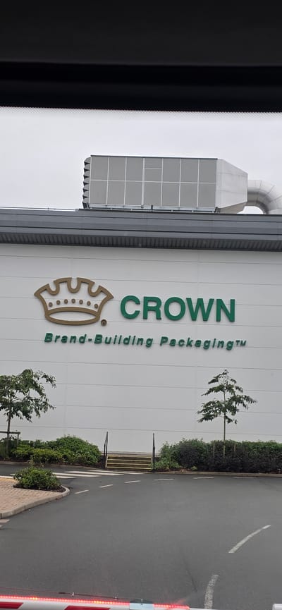 The image shows the exterior of the Crown Packaging building, located in Peterborough, United Kingdom. The building features a large white facade with the company's logo, a golden crown, and the word "CROWN" in green, accompanied by the tagline "Brand-Building Packaging™". Above the facade, there is a large grey ventilation unit. In front of the building, there are two young trees and a line of green bushes. A paved driveway with white markings leads to the building, with a pedestrian crossing and a set of yellow-striped stairs with black handrails. The sky is overcast and grey. The lower part of the image shows a red and white striped barrier, suggesting an entrance or exit to the property.