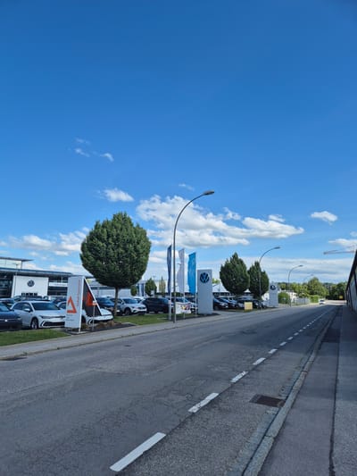 The image depicts a street scene in Landshut, Germany, likely in a commercial area given the presence of a Volkswagen dealership. The sky is a vibrant blue with scattered white clouds. In the foreground, a two-lane asphalt road runs from the lower left to the upper right of the frame. The road surface shows signs of wear and tear with visible cracks and patches. White dashed lines delineate the lanes. To the right of the road, a concrete sidewalk is visible, bordered by a stone curb. A metal grate is set into the road near the curb.
On the left side of the road, a Volkswagen dealership is apparent. Several Volkswagen cars are parked in rows on a landscaped area in front of the building, which features a modern architectural design with large windows. A prominent sign displays the Volkswagen logo, and another sign reads "Unfall Spezialist" (Accident Specialist) with additional text in smaller font indicating "Karosserie," "Lack," and "Clever Repair." Two tall, cone-shaped evergreen trees stand near the dealership. Blue and white flags, also featuring the Volkswagen logo, are displayed on poles. Streetlights are positioned along the road.
Further down the street, more trees and buildings are visible, suggesting a suburban or industrial edge of the city. In the distance, a yellow construction crane can be seen against the sky. The overall impression is a bright, sunny day at a car dealership on a city street. No people are visible in the image.