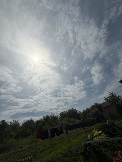 In Septemvritsi, Bulgaria, an outdoor scene unfolds under a partly cloudy sky with the sun visible and bright. The foreground features a clothesline laden with dark-colored garments, including a red item, hanging to dry. Beyond the clothesline, lush green grass and dense foliage dominate the landscape, with trees forming a thick border. Partially visible in the background is a rustic brick house with a tiled roof, nestled amongst the trees. The overall impression is a rural, domestic setting in a village environment.