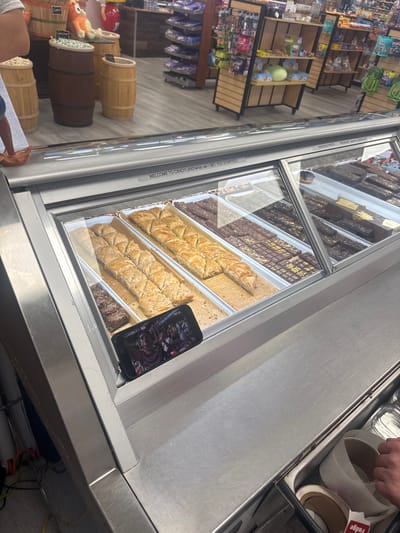 In a candy store in Ocean City, United States of America, a refrigerated display case is filled with various confectionery items. The display features rows of baklava, chocolate bars, and other sweets. A smartphone is propped up on the edge of the display case, showing a live video feed of a sports event, with several individuals visible on the screen. In the foreground, a person's hand is seen reaching into a bin containing rolls of tape. The background reveals shelves stocked with more candy and merchandise, suggesting a busy retail environment.