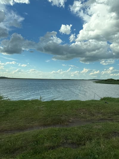 This image, captured in Айдарлы, Kazakhstan, presents a wide-angle view of a large body of water, likely a lake or reservoir, under a dynamic sky. The upper half of the frame is dominated by a blue sky interspersed with significant cloud cover, ranging from bright white, fluffy cumulus clouds to darker, denser stratocumulus formations. The water surface, occupying the mid-ground, exhibits visible ripples and texture, indicating the presence of wind.
Along the distant left horizon, a series of white wind turbines are clearly visible, suggesting industrial or renewable energy infrastructure. Towards the right side of the water, the shoreline curves, revealing a natural bank heavily populated with green reeds and other vegetation, with a low hill visible in the far distance beyond it.
The immediate foreground consists of a gently sloping grassy bank leading down to the water's edge. This bank is covered in vibrant green grass, interspersed with patches of drier vegetation and a visible dirt path or worn track running horizontally across the lower part of the frame. On the right side of this grassy bank, two fishing rods are set up on tripods, positioned to cast into the water. A blue bucket or container is resting near the base of the fishing equipment. No people are visible in the scene. The overall lighting suggests natural daylight conditions.