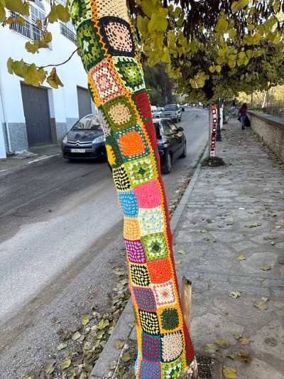 The image depicts a street scene in Setenil de las Bodegas, Spain, characterized by a yarn-bombed tree trunk in the foreground. The prominent subject is a tree trunk wrapped entirely in a vibrant, multicolored crocheted "granny square" blanket, featuring a patchwork of green, yellow, orange, red, pink, blue, beige, brown, and black squares stitched together. The tree, displaying yellowish-green foliage, stands next to a gray asphalt road on its left and a stone-paved pedestrian pathway on its right.
To the left of the main tree, a dark blue Citroën C4 Picasso with Spanish license plate 3394 FZG is parked partially on the road, adjacent to a white building featuring a dark gray garage door and a white balcony on the upper level. Other vehicles are visible further down the street. The road surface has scattered brown and yellow leaves near the curb.
To the right, the irregular stone-slab pathway, also strewn with fallen leaves, extends into the distance. Another smaller tree trunk further along this pathway is similarly adorned with a crocheted wrap, primarily in pink and white squares. A low stone wall with a reddish-brown top edge runs along the far right side of the pathway. Two indistinct figures are visible walking away on the pathway in the distance, one wearing dark clothing and appearing to carry an object in their left hand. The scene is illuminated by natural daylight.