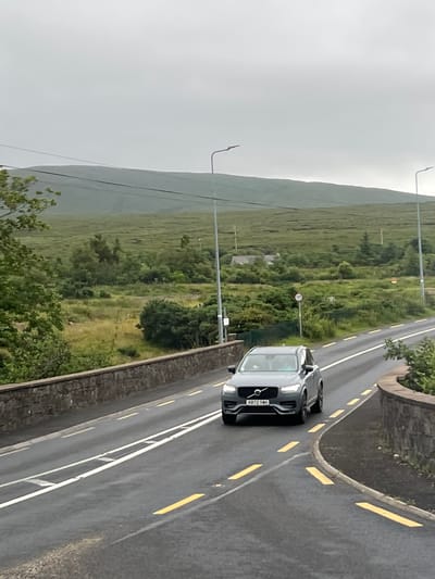 The image shows a dark gray Volvo SUV driving on a wet asphalt road, likely in Gweedore, Ireland. The car is positioned on the right side of the road, with its front facing towards the viewer. The license plate is visible and reads "KR72 YXH". The road has yellow and white markings.
The scene is a rural landscape with rolling green hills covered in lush vegetation. The sky is overcast and appears to be a light gray, suggesting it is either early morning or a cloudy day. The road is bordered by a stone wall on the right, and beyond that, there is more greenery and trees. On the left side of the road, the terrain slopes upwards, covered in dense green grass and some scattered trees, extending to a misty mountain in the background.
A speed limit sign showing "100" is visible on the side of the road in the middle ground, indicating a 100 km/h speed limit. There are also lampposts on the side of the road. The overall mood of the image is calm and perhaps a bit somber due to the overcast weather. The wet road surface suggests recent rain or ongoing light precipitation.