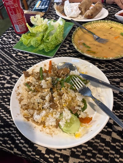 The image captures a tabletop dining scene in Nongkhiao-Nua, Laos, likely post-meal. In the foreground, a white oval plate displays remnants of fried rice, containing white rice grains, brown meat pieces, green beans, orange carrot fragments, and a cucumber slice. A silver spoon and fork rest on the plate. To the left, a green rectangular plate holds several fresh green lettuce leaves. Behind it, a red 325 ml Coca-Cola can, bearing Lao text, stands upright. A dark-colored smartphone with an illuminated screen displaying numerical digits is positioned behind the soda can. To the right, a round, shallow bamboo-weaved plate with a green rim contains a small amount of pale yellow sauce, a single silver fork, and a few pieces of green beans and carrots. In the upper right background, a white round plate holds two partially consumed spring rolls and a crumpled white napkin. A human hand with a red wristband is visible in the top right corner. The table is covered by a black and white geometric patterned tablecloth.