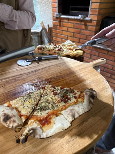 The image depicts an indoor or semi-enclosed culinary space in Porto Alegre, Brazil, focused on pizza preparation and serving. In the foreground, a large, circular wooden pizza peel or serving board displays multiple slices of thin-crust pizza. Several slices are topped with melted cheese and dried green herbs. Other slices also feature small, diced red ingredients, possibly meat or sun-dried tomatoes. One pizza segment on the board appears partially consumed. A metal pizza cutter with a black handle and a reflective metallic cylindrical object rest on the board's left side. In the mid-ground, a hand from the right side of the frame, holding a metal pizza server, suspends a freshly baked, uncut pizza or a large segment above the board. This suspended pizza also exhibits melted cheese and dried herbs. The background reveals a prominent brick oven, constructed from reddish-brown bricks with a dark, open interior. To the left of the oven, a light-colored wall is visible, with hints of other household or kitchen elements in soft focus. Partial views of two individuals are present: the upper torso of one person, wearing a light pink long-sleeved shirt under a tan vest, is visible on the left, while the hand holding the pizza server belongs to another individual on the right.