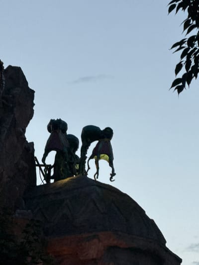 The image depicts a silhouette of figures atop a rocky structure against a pale sky. The figures appear to be stylized, possibly statues or puppets, with multiple limbs and flowing, cape-like elements. Their forms are dark against the lighter background. To the upper right, a portion of dark green foliage is visible, also in silhouette. The overall scene suggests an outdoor setting, likely during twilight or early morning. The image does not contain sufficient information to ascertain a specific location in Vienna, Austria.