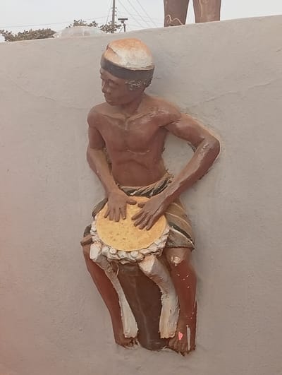 A 3D sculpture of a muscular, dark-skinned man playing a round drum. He wears a head wrap and a striped loincloth. His hands are positioned on the drum's surface. The sculpture is mounted on a light-colored wall, seemingly outdoors. Above and partially behind the figure, there are other sculpted forms and a distant view of utility poles and trees. The lighting is soft, creating subtle shadows that highlight the figure's contours.
