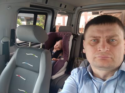 The image shows the interior of a car, likely a van. In the foreground, a man wearing a blue striped button-down shirt and earbuds is looking at the camera. In the background, a baby is asleep in a purple car seat. The car appears to be parked, as indicated by the lighting and the visible scenery outside the windows, which suggests a residential or commercial area. The overall scene depicts a moment of travel with a child. The location of Altdorf bei Nürnberg, Germany is not determinable from the visual information in the image.