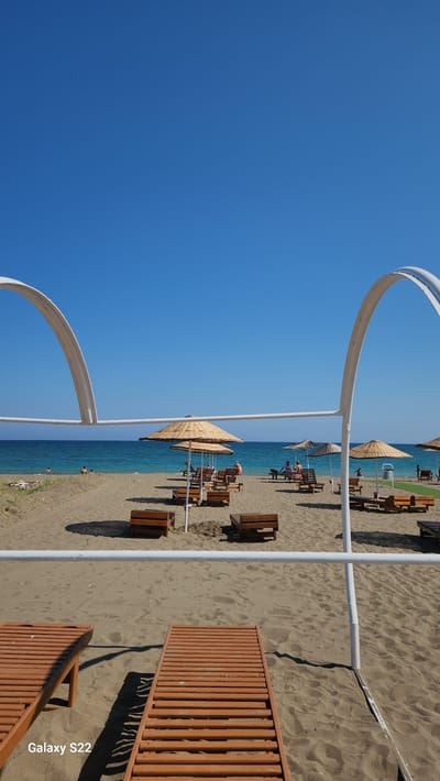 This image depicts a sunny beach scene in Erdemli, Turkey, viewed from a low angle on the sand. The foreground features two partially visible brown wooden sun loungers on textured light-brown sand. A white architectural structure consisting of two curved arches connected by a horizontal bar frames the middle ground. The middle ground presents a sandy beach furnished with numerous straw-thatched beach umbrellas and corresponding wooden sun loungers and small benches arranged in rows. Several individuals are present: two people are in the turquoise-blue sea on the left, while others are seen lounging on sunbeds or sitting on benches on the sand, primarily in the central and right portions of the frame. Some small patches of green grass and a white refuse bin are visible on the far right side of the beach. The background features the calm, deep-blue Mediterranean Sea stretching to the horizon, where a distant landmass is faintly visible on the right. The sky above is clear and intensely blue, devoid of clouds. A "Galaxy S22" watermark is present in the bottom left corner of the image.