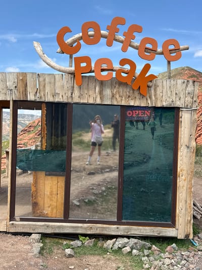 The image shows a sign for "Coffee Peak" made of orange letters attached to a wooden frame, with a glass window behind it. The window reflects the surrounding environment, including a dirt path, people walking, and a mountain in the distance. An "OPEN" sign is visible in the window. The structure is located outdoors, with a rocky and grassy area to the right. Based on the provided context, this scene likely exists in Jeti-Ögüz, Kyrgyzstan.