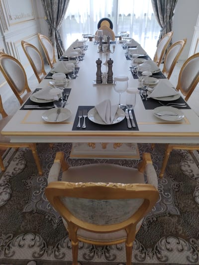 A formal dining room is depicted with a long, white, rectangular table set for a meal. The table is adorned with white napkins folded neatly on plates, silverware, wine glasses, and water glasses. Two salt and pepper grinders stand in the center of the table. The chairs surrounding the table are upholstered in a light-colored fabric and feature ornate gold-painted frames, matching the gilded trim on the table and the elaborate wall paneling. Heavy, patterned curtains hang from a large window at the far end of the room. The floor is covered with a large, ornate rug with a dark gray and cream pattern. The overall impression is one of opulence and formality. The image is identified as being located in the city of Konigil, Uzbekistan.