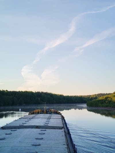 The image captures a serene view from the stern of a large, flat-topped barge or cargo ship as it navigates a calm, wide river or canal. The vessel dominates the foreground, with its grey deck visible, punctuated by numerous circular hatch covers. A yellow structure with machinery is situated towards the rear of the deck.
The sky is a pale, clear blue, adorned with two prominent, wispy white contrails that cross each other in the upper portion of the frame. Below the horizon, dense green forests line both banks of the waterway, their reflections visible on the glassy surface of the water. A subtle mist or haze hovers over the distant water.
The sun, likely low in the sky during either early morning or late afternoon, casts a gentle, golden light on the distant treetops, particularly on the right bank. The barge is clearly in motion, evidenced by the distinct V-shaped wake trailing behind it on the right side of the frame. A white, diamond-shaped navigational buoy floats in the river channel on the left. The scene evokes a sense of peaceful travel through a natural landscape. No discernible text is visible.