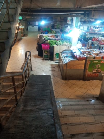 An indoor market with a partially visible staircase leading to the lower level. Produce displays are present, with a woman in a hijab browsing. A vendor stands behind the vegetable counter. A freezer cabinet and promotional signage are visible nearby. The flooring is tiled. The lighting appears to be a mix of fluorescent and natural. The scene seems bustling and utilitarian.