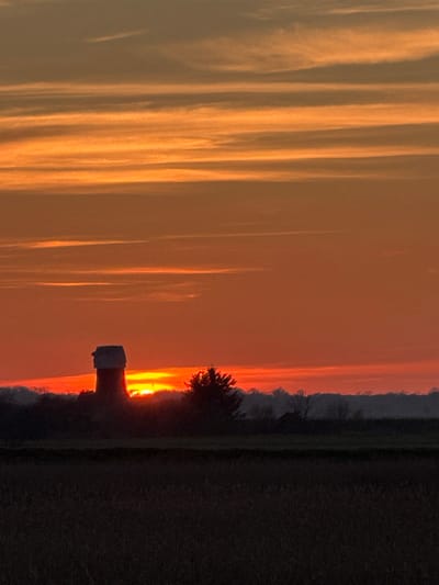 The scene depicts a rural landscape at either sunrise or sunset within Broadland, United Kingdom, consistent with the Norfolk region. A prominent dark silhouette of a brick tower windmill, featuring a lighter cap, stands on the left midground horizon. The sun, a brilliant yellow-orange orb, is partially obscured directly behind the windmill and a cluster of dark, possibly coniferous, trees positioned immediately to its right. The sky exhibits vibrant orange and red stratifications with horizontal cloud formations. A faint line of distant deciduous trees forms the far horizon, with a small, dark, triangular-roofed building silhouette visible to the far right. The foreground consists of a dark, uncultivated agricultural field. No human figures, actions, or interactions are discernible. The dramatic backlighting creates deep silhouettes against the intensely colored sky.