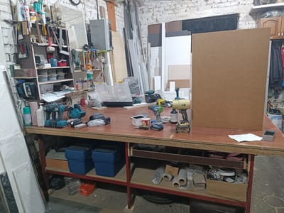 The image depicts a workshop setting, likely in Pokoinoe, Russia, characterized by a cluttered workbench filled with various tools and materials. The workbench itself is constructed from wood and metal, with shelves underneath storing more items. On the surface, several power drills, batteries, screws, and other hardware are visible. A large, light brown particle board stands upright on the right side of the workbench, dominating that portion of the scene. The background reveals a brick wall, shelving units stocked with supplies, and various construction materials leaning against the wall. The overall impression is one of an active workspace, possibly for woodworking or general repairs. There are no people in the image.