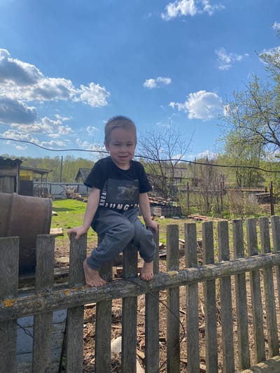 A young boy with short blond hair is perched barefoot on a weathered wooden fence. He wears a dark t-shirt and grey pants. The backdrop consists of a partly cloudy sky and rural scenery, featuring trees, grass, and structures including a metal barrel and a building with a yellow tarp visible. The image suggests an outdoor setting, possibly a yard or countryside area. The location mentioned, Узяр, Russia, cannot be definitively determined from this image alone.