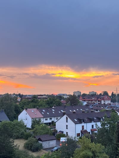 This image depicts a residential area in Sindelfingen, Germany, at dusk. The sky is filled with dramatic clouds illuminated by the setting sun, casting an orange and yellow glow. In the foreground, several houses with red-tiled roofs are visible, surrounded by lush green trees. A small, concrete-block outbuilding is situated in the lower left portion of the image, next to a patch of dry grass. A dark-colored car is parked near the outbuilding. In the background, taller buildings and a cell tower are visible, indicating a developed urban or suburban setting. The overall atmosphere is serene and picturesque, characterized by the interplay of natural light and human-made structures.