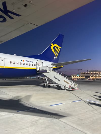 The image shows a Ryanair Boeing 737-800, registration EI-IGK, parked at Malta International Airport in Luqa, Malta. A mobile stairway from "Aviaserve" is attached to the aircraft's rear door. The tail fin of the aircraft is visible, displaying the Ryanair logo. The airport terminal and other structures are visible in the background. The sky is clear and blue. The ground surface is concrete with painted lines. Shadows suggest it is either early morning or late afternoon. There are no people visible in the image.