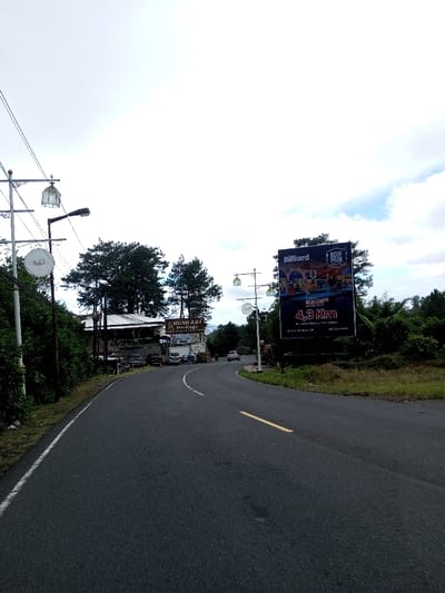 This image captures a two-lane asphalt road curving gently to the right under an overcast sky in the city of Phoxay, Laos. The road surface is dark gray, featuring a solid white line on its left edge and a dashed yellow line in the center. Dense green vegetation, including tall coniferous trees, borders both sides of the road.

On the left side, a series of ornate white street light poles, each with a lantern-style fixture and a circular white sign, line the roadside. Behind these poles and vegetation, a complex of white-roofed buildings is visible, appearing to be commercial establishments. Prominent signs on these buildings include "BUMIAKI HERITAGE" and "ONE TEO." Several vehicles, including a white van, are parked or stationary near these buildings.

On the right side of the road, further into the scene, a large dark-colored billboard stands amidst green foliage. The billboard advertises "Billiard" and "ROOF PARK," displaying images of a pool table and an outdoor leisure area, along with the text "4,3 Km" indicating a distance. Similar ornate white street light poles are also visible on this side of the road, extending into the distance. Two white vehicles are present further down the road, traveling away from the camera. No individuals are clearly discernible.
