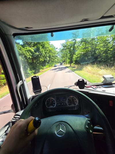 The image shows the interior of a truck cab, looking forward through the windshield. The driver's hands are visible, holding a yellow object with a black tip, possibly a vape pen or a small tool. The truck is on a paved road, bordered by a grassy verge and trees on both sides. The road is straight and appears to be in a rural or semi-rural area. In the distance, other vehicles can be seen on the road. The dashboard of the truck is visible, with a steering wheel, speedometer, and various other gauges and controls. A smartphone is mounted on the windshield. The sky is blue with some clouds, and the sun is shining, indicating a bright, clear day. There is no direct indication of the location being in Wittenberg, Germany, but the image depicts a typical scene of road travel in a European setting.
