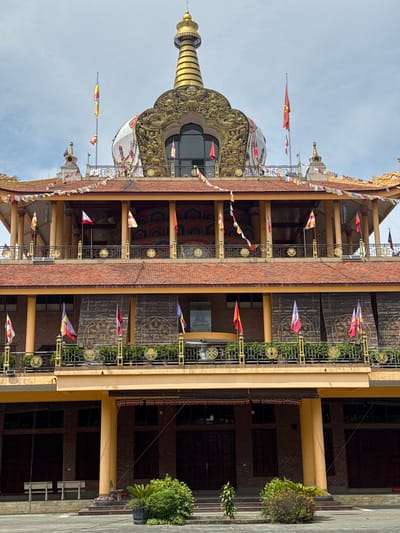 The image shows the exterior of a Buddhist temple in Việt Trì, Vietnam. The temple features traditional East Asian architectural elements, including a tiered roof, ornate decorations, and prayer flags. The central focus is a large, golden stupa at the top of the structure, surrounded by smaller stupas and decorative motifs. The building is constructed with brick and painted in warm tones of orange and yellow. Prayer flags in various colors are strung across the balconies and roofline. The foreground shows a paved courtyard with some greenery, including potted plants and bushes. The sky is overcast. There are no people visible in the image.