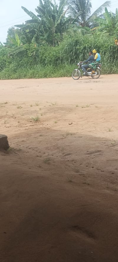 The scene depicts a dirt road in Amauzu, Nigeria. Two people are riding a motorcycle on the road. The area is lined with dense green vegetation, including banana trees. The foreground is shaded.