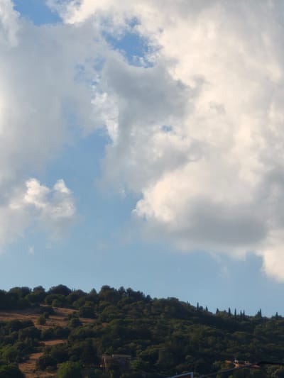 The image shows a rolling, tree-covered hillside with a bright blue sky dotted with fluffy white and grey clouds above. In the distance, on the crest of the hill, a line of tall, slender cypress trees stands out against the sky. A few buildings are visible nestled among the trees on the lower slopes of the hill. The vegetation is a mix of dark green trees and lighter brown, dry grass. The overall impression is one of a peaceful, natural landscape in a rural setting. This scene is representative of the landscape in and around Chiugiana, Italy.
