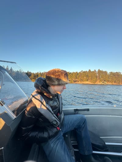 A person is seated in a metallic grey motorboat on a body of water near Boda, Sweden. The individual, positioned off-center left, wears a brown fur trapper hat, a shiny black puffer vest over a grey long-sleeved garment, blue jeans, and dark footwear. A red safety clip is visible on the person's chest. The individual is oriented towards the right side of the frame, with their head slightly lowered, looking away from the camera's direct line of sight. The boat's clear windshield on the port side exhibits water droplets. The background consists of a forested shoreline with numerous coniferous trees, illuminated by low-angle sunlight. A small light-colored building with a dark roof and a short dock is visible on the far right of the distant shoreline. The water surface shows ripples, reflecting the clear blue sky.