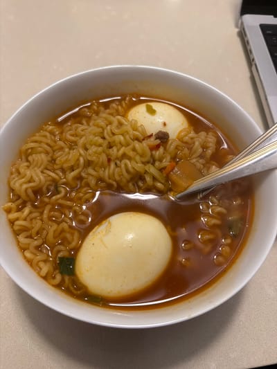 In a white ceramic bowl, ramen noodles are submerged in a reddish-brown broth. Two halved boiled eggs are visible, one on top of the noodles and the other submerged in the broth. A fork is partially submerged in the bowl, resting on the noodles and broth. The bowl is placed on a light-colored, textured surface, possibly a countertop, with a portion of a laptop visible in the upper right corner. The scene appears to be indoors, likely a home or office environment.