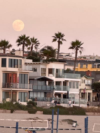 This image captures a twilight scene in Manhattan Beach, California, USA, focusing on a row of multi-story beach houses. A large, pale moon is visible in the upper left quadrant of the sky, appearing soft-edged and slightly hazy against a pale pinkish-orange sky. Several palm trees, characteristic of the coastal environment, are scattered throughout the frame, with their fronds silhouetted against the sky.
The beach houses exhibit a modern architectural style, featuring balconies with glass railings, large windows, and a mix of light-colored stucco and siding. Some houses have rooftop patios or balconies. There are visible elements of outdoor living, such as patio furniture, umbrellas, and vegetation like bushes and grasses.
In the foreground, a beach volleyball net is set up, suggesting recreational activity. Several people are visible on the elevated walkway or terraces of the beach houses, some walking, others standing and conversing. One person is seen near the volleyball net, holding a paddleboard. The overall impression is a serene, coastal residential setting at dusk.