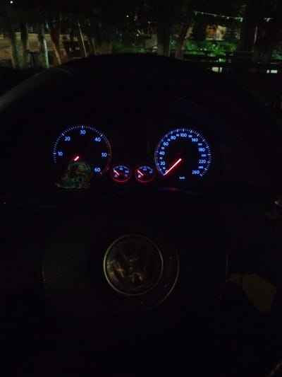 The image shows the illuminated dashboard of a Volkswagen car at night. The speedometer reads approximately 25 km/h. The fuel gauge indicates the tank is slightly above half full, and the temperature gauge appears to be at a normal operating temperature. The location of Bazan, Bulgaria, cannot be determined from the image. The steering wheel, with the Volkswagen logo, is in the foreground. The background is dark, with some blurry lights and shapes suggesting trees or buildings.