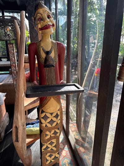 An indoor scene in Tibubiyu, Indonesia, displays a prominently featured, intricately carved wooden statue standing upright. The statue, appearing to be a traditional Indonesian figure or puppet, has a light-colored face with red lips, black and red eyes, and a dark, pointed hairstyle. It wears a red upper garment with gold trim and a black, carved necklace with a pendant. The lower body is pale yellow with dark brown carved geometric patterns. The statue holds a rectangular, shallow, dark wooden tray with carved edges at chest height. To the statue's left, a tall, light-colored wooden carving leans against an unseen support, and a woven rattan light fixture hangs from the ceiling further back. The floor transitions from dark wood on the left to patterned tiles (blue, orange, cream) on the right. The background consists of tall glass windows with dark wooden frames, allowing bright natural light into the space and revealing lush green tropical foliage outdoors, along with a gravelly ground surface and a faint red and yellow object in the distance.