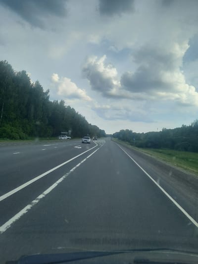 This media file captures a view from a vehicle traveling on a highway in Petrovka, Russia. The scene is dominated by a paved road with white lane markings, stretching into the distance. On the left side of the road, a dense forest of green trees lines the highway. The sky above is partially covered with dramatic, grey and white clouds, suggesting a changeable weather pattern. A few vehicles are visible on the road, including a white car further ahead and another car seemingly stopped on the shoulder, partially obscured by trees. There are also road signs visible along the side of the road, though the text on them is not legible. The perspective suggests the photo was taken from the passenger seat of a moving car. The lighting indicates it is daytime, possibly late afternoon or early evening, given the softness of the light and the contrast in the clouds.
