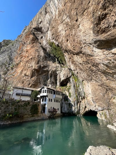 The image displays the Blagaj Tekke complex situated at the base of a significant rock formation in Dračevice, Bosnia and Herzegovina. The scene features a multi-story white building, characterized by dark window frames and roof accents, built partially into the base of a towering, striated limestone cliff and extending over a body of tranquil, turquoise water. A wooden balcony on the building's lower level accommodates two discernible individuals standing. Adjacent to the main structure on the left, a connected white building rests on a stone retaining wall. The colossal cliff, composed of light brown and gray rock, exhibits vertical striations and is partially covered with sparse green vegetation, including moss and small plants, particularly near a water seep on the right side. A large, dark cave entrance is visible at the cliff's base on the right, where the water appears to originate. The sky is clear blue overhead, and reflections of the architecture and cliff are present on the water's surface.