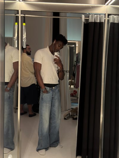 A Black man wearing a white short-sleeved shirt and baggy blue jeans stands in a fitting room and takes a selfie in a mirror. He has white earbuds in his ears and is holding a smartphone. His left hand is in his pocket, and his right hand is holding the phone. A man in a yellow shirt and dark shorts is reflected in the mirror behind him. The fitting room has a black curtain to the right and a white curtain to the left. The floor is white, and there are clothes on a rack in the background. The location is identified as Fibracolor, Spain.