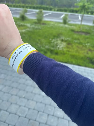 The image shows a close-up of a person's left arm and wrist, wearing a dark blue sweater and a white wristband with yellow trim. The wristband has text printed on it in black, which appears to be in Russian. The visible text on the wristband reads: "Дата рождения: 16.09.2007" (Date of birth: 16.09.2007), "Пациент: РУСАКОВА И.В." (Patient: RUSAKOVA I.V.), and "Номер ИВ:" (IV number:). The background is out of focus, showing a green grassy area with some white flowers and what appears to be a paved area with parking lines. The scene is likely outdoors, possibly in a park or recreational area. While the image does not explicitly show the city of Solnechnoye, Russia, the presence of Russian text on the wristband suggests a possible connection to this location.