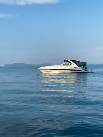 The image shows a white yacht with a black canopy, anchored in calm blue water under a clear sky. The yacht is positioned vertically in the frame, with its bow pointing upwards. A person can be seen standing on the deck of the yacht, near the stern, appearing to be engaged in an activity. The background reveals a hazy coastline with distant mountains and a few other boats, suggesting a summer day and a coastal setting. The water is gently rippling, reflecting the sunlight. The overall mood is peaceful and serene, characteristic of a day spent out on the water. The time of day appears to be late morning or early afternoon, given the brightness of the sky and the quality of light. The location is Milvignes, Switzerland, as indicated by the context, implying a freshwater lake environment. No visible text is present in the image.