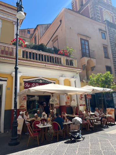 The image shows a street scene in Sorrento, Italy, on a sunny day. A yellow building with a balcony decorated with red flowers and potted plants is visible on the left, with an ornate lamp post in the foreground. In the center, an outdoor cafe with white umbrellas is set up on a cobblestone street. Several people are seated at tables, eating and conversing. The cafe appears to be a popular spot, with signs advertising "Gelateria" and "Breakfast." The architecture suggests a historic European city, and the presence of citrus trees and the specific style of the buildings point to the Campania region of Italy.