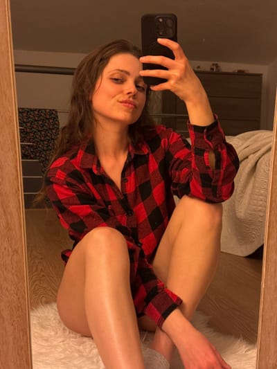 A female subject is depicted in a full-length mirror selfie, positioned sitting on a white fluffy rug over a wooden floor in an indoor environment in Cava de' Tirreni, Italy. She has long, wavy brown hair and is wearing an unbuttoned red and black buffalo plaid shirt, which reveals her bare legs. Her left hand holds a dark-colored smartphone, equipped with a pop socket, capturing the image reflected in the mirror. Her right arm is bent, with the forearm resting across her lap. She is seated with her legs crossed; her left foot wears a light grey sock while her right foot is bare. The subject is looking directly into the mirror with a slight smirk. The background, visible in the mirror's reflection, includes a dark wooden dresser with multiple drawers, a light-colored quilted blanket or comforter draped over an unseen surface, and a patterned garment hanging on a rail to the left. The room is illuminated with warm lighting.