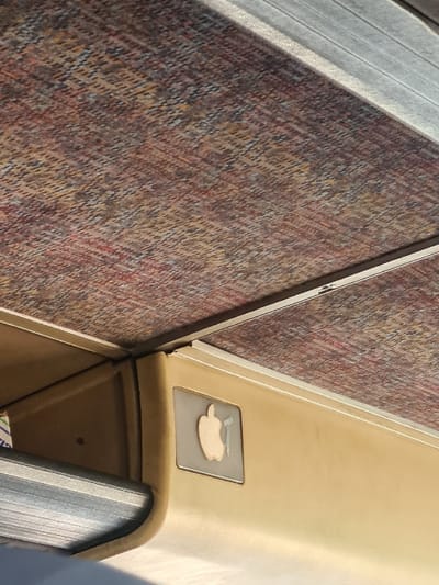 The image displays the interior of a vehicle, viewed from a low angle, focusing on the ceiling and upper side panel. The ceiling is covered with a textile panel featuring a textured, diagonal pattern predominantly in shades of red, brown, and beige. Below this, a smooth, light beige-tan panel runs horizontally, possibly representing an upper wall or bulkhead. Attached to this beige panel is a rectangular sign depicting a light-colored apple silhouette positioned next to a smaller, stylized blue-green object resembling a pipe or handle. A metallic-gray, ribbed surface, likely the underside of an overhead luggage rack, is partially visible in the lower-left quadrant. No individuals or specific identifying markers for the city of Al-Kiswa, Syria, are present within the frame.