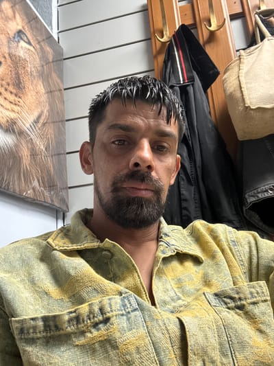 The image is a close-up, waist-up, selfie-style portrait of a man in a yellow and blue patterned shirt. The man has dark, wet-looking hair, a beard, and appears to be in his late 30s or early 40s. He is looking directly at the camera with a neutral expression. Behind him, there is a wall with horizontal white slats. To his left, a portion of a large framed image featuring a lion's face is visible. To his right, a wooden coat rack with several hooks is mounted on the wall. A black jacket with red and white stripes on the collar and a beige canvas bag are hanging on the coat rack. The man is positioned in the foreground, with the background slightly out of focus. The lighting suggests an indoor environment. There are no discernible landmarks that would indicate a specific location within Nuremberg, Germany.