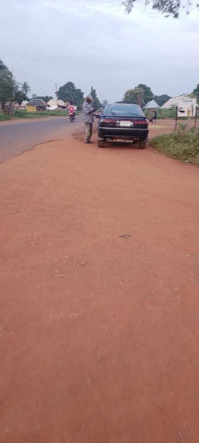 The image depicts a roadside scene in Ichol, Nigeria, under an overcast sky. A dark-colored sedan is parked on the right side of a dirt road, facing away from the viewer. A person wearing a plaid shirt and khaki pants stands beside the driver's side of the car, appearing to interact with something near the vehicle. In the background, a red motorcycle is visible on the paved road to the left, with a rider on it. Further down the paved road, several small buildings with corrugated metal roofs are scattered among trees. A white sign with text and graphics is mounted on a post on the right side of the road. The ground is predominantly red dirt, with patches of dry grass and vegetation along the road. The overall environment suggests a rural or semi-urban setting.