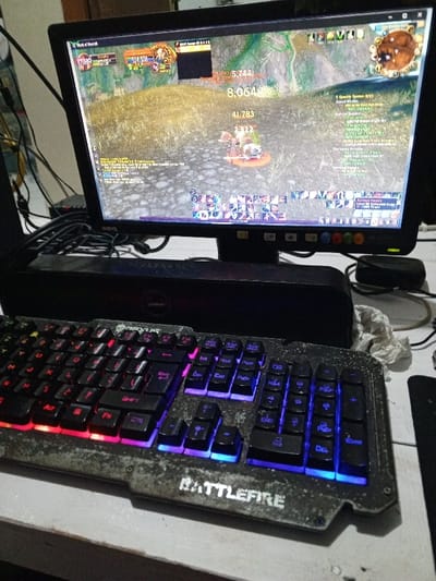 This image depicts a gaming computer setup, located in the city of Al Haouafate, Morocco. The central focus is a black BenQ LCD monitor displaying a complex user interface from a fantasy-themed Massively Multiplayer Online Role-Playing Game (MMORPG), likely World of Warcraft. The game scene shows an outdoor environment with a character engaged in combat against a target on a grassy, textured ground, indicated by large damage numbers "8,064" and "41,783" floating above. Various game UI elements are visible, including health and mana bars, a mini-map, quest logs, chat window, and multiple rows of action bar icons.

Below the monitor, a black "JURASSIC" branded soundbar or speaker is positioned horizontally. In front of the speaker rests a heavily used "BATTLEFIRE" branded gaming keyboard. The keyboard features distinct RGB backlighting, with the left key cluster (WASD and surrounding keys) illuminated in shades of red, pink, and purple, while the right key cluster (arrow keys, numpad, and function keys) displays blue and light blue illumination. The keyboard's surface exhibits a mottled, textured appearance consistent with wear or design.

The entire setup rests on a light-colored, possibly white or off-white, desk surface that shows some minor scuffs or discoloration. To the left of the monitor, a tangle of black cables is visible. To the right of the speaker, a small, black, oval-shaped peripheral (possibly a mouse) and a crumpled piece of white paper or tissue are present. The background behind the monitor is an indistinct light-colored wall. No individuals are visible in the scene.