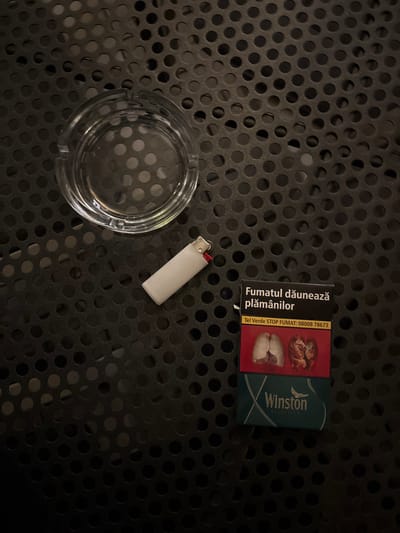 An ashtray, a white lighter, and a pack of Winston cigarettes rest on a black, perforated metal surface. The cigarette pack features a health warning with images of damaged lungs. The ashtray is clear and empty. The lighter is a common disposable type. The scene suggests a smoker's setup, possibly outdoors, given the metal surface.